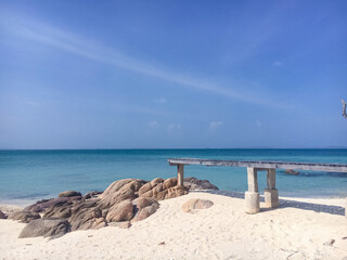 Fototapeta premium A wooden pier extending over a pristine white sand beach with turquoise sea water under a clear blue sky.