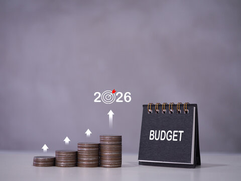 A small black desk calendar displaying the word 'BUDGET' next to growing stacks of coins with upward arrows leading to a 2026 target