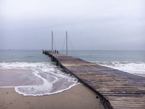 A wooden pier extending from the sandy beach into the sea as gentle waves create white foam on the sand under an overcast, cloudy sky.