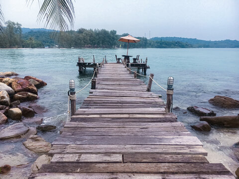 A wooden boardwalk stretching into a calm bay, with rocky shores on the side and a lush green island in the background
