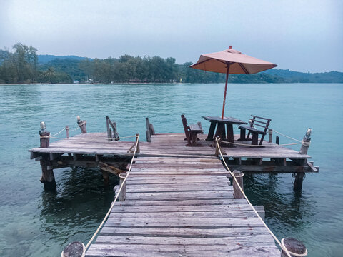 An outdoor wooden deck on a pier over the sea, featuring a dining table, wooden benches, and a large sun umbrella
