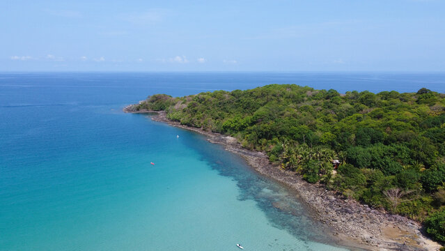 An aerial drone photograph of a lush tropical island with dense green forest and a rocky shoreline meeting the clear blue ocean