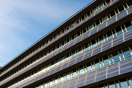 Solar panels line a long building facade under blue sky, while windows, balconies and railing draw urban architecture into perspective