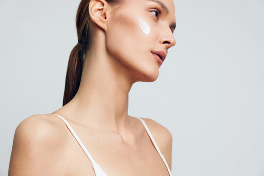 Young woman with clear skin applying white face cream on cheek. Side profile on light gray background. Concept of skincare, beauty routine, and healthy skin care. Soft natural lighting.