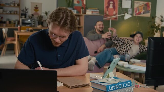Caucasian young adult male student trying to study at desk while distracted by noisy roommates watching TV in background.Commercial videos about student life challenges and shared housing