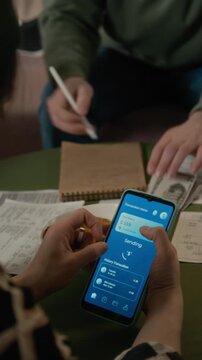 Vertical of young zoomers using mobile banking app on smartphone to transfer money while man counting cash at table in student dorm. Promoting digital payment systems and personal finance apps