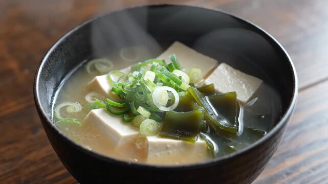 Steaming hot Japanese miso soup with tofu cubes wakame seaweed and green onions in a black bowl