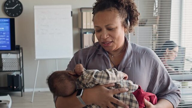 Portrait of middle aged African American woman wearing headset, holding sleeping baby while working in office. Clip is perfect for marketing remote work, work-life balance, and hybrid employment
