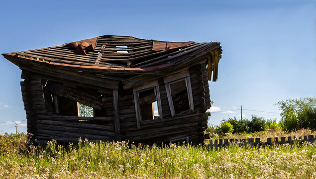 Abandoned wooden house in rural Russian village landscape during summer. Ruined countryside building standing in grassy field under blue sky, concept of rural decline and depopulation