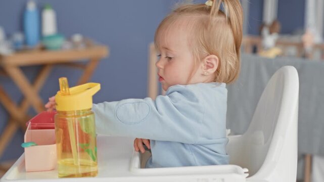 Girl toddler leans in highchair and reaches with hand for yellow plastic bottle on tray beside a pink container inside building; curiosity.