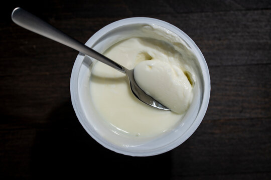 White creamy yogurt in plastic cup with a spoon on wooden background. Top view of thick organic yogurt or sour cream inside an opened plastic container with a metal spoon on dark wooden table