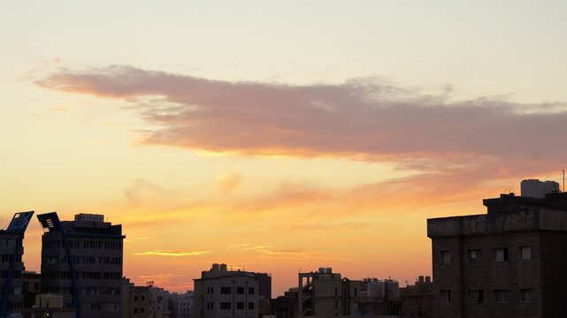 A wide static shot of a beautiful orange and yellow sunset sky with soft clouds over the dark silhouettes of urban residential buildings in Kuwait.