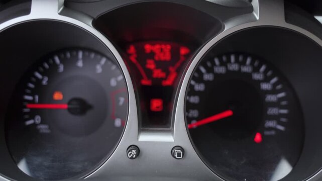 A close-up shot of an illuminated car dashboard display, showing a glowing speedometer, tachometer dials, and a red digital center screen while stationary.