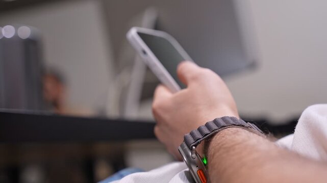 A close-up shot of a man wearing a modern smartwatch while holding an iPhone in an office or studio setting with a shallow depth of field.