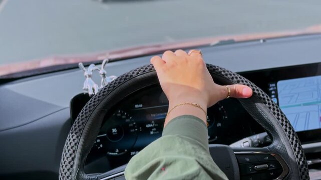 A close-up POV shot of a woman's hand gripping a textured steering wheel while driving a modern car. The digital dashboard is visible as she navigates behind another vehicle on a sunny daytime road.