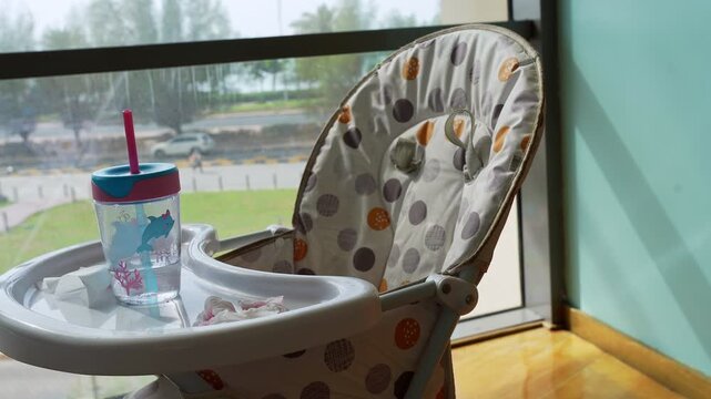 Empty baby high chair with a colorful sippy cup and tissues on the tray, sitting indoors by a large glass window. Bright natural daylight illuminates the scene with a view of a busy city street.