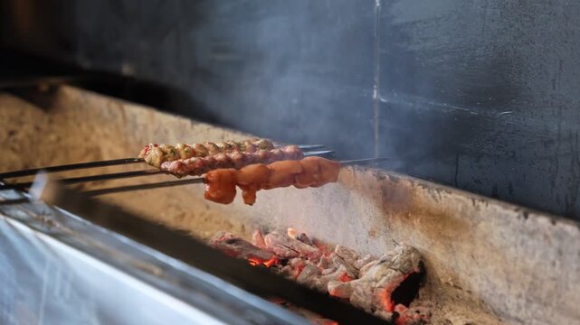 A close-up shot of traditional meat and chicken skewers roasting over a hot smoking charcoal pit. The delicious Middle Eastern kebabs cook slowly on metal rods in a dark, moody kitchen setting.