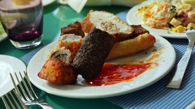 A close-up shot of a mixed buffet appetizer plate on a blue placemat at a restaurant table. The white dish holds sushi, kofta, fried snacks, and sweet chili sauce during a bright daytime meal.