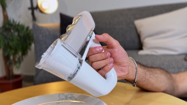 a man holding a white electric vegetable slicer over a plate. The handheld kitchen appliance rests above a wooden table indoors, featuring a shallow depth of field with a blurred sofa.