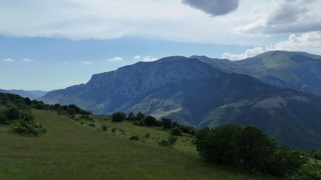 Paesaggio montano con dolci colline erbose in primo piano e una cresta rocciosa illuminata dal sole sullo sfondo. Cielo parzialmente nuvoloso e atmosfera ampia, naturale e luminosa