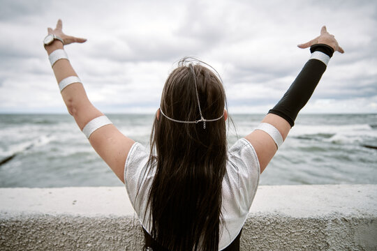 Young woman cosplay with long dark hair in silver costume raising arms by ocean under cloudy sky