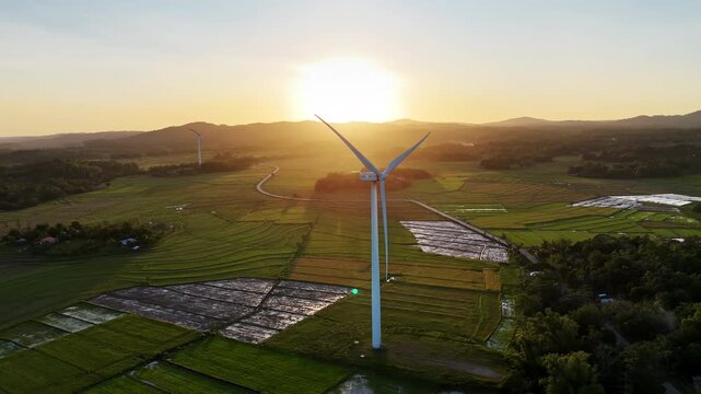 Aerial drone view of a white wind turbine silhouetted against a golden sunset sky above patchwork green and flooded rice paddies, San Lorenzo Wind Farm, Philippines