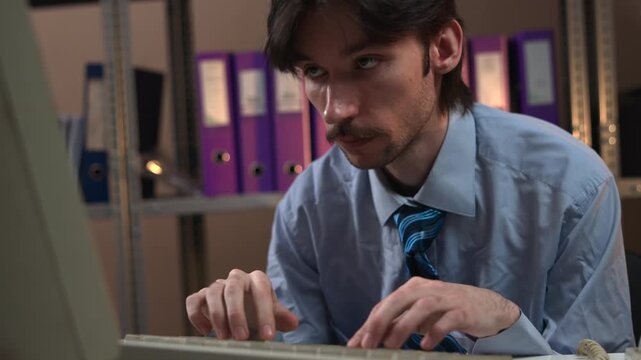 Focused man working on laptop at office desk with binders and shelves in the background