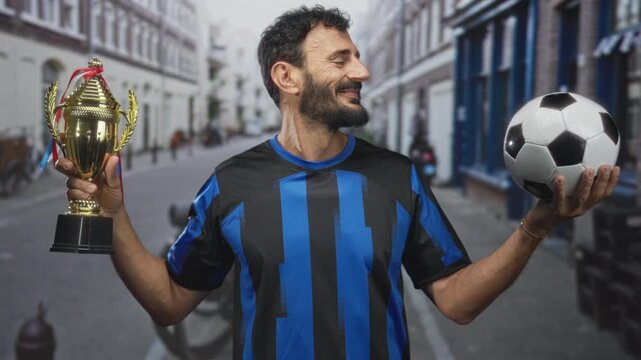 Man in blue black soccer uniform holds trophy in left hand and a soccer ball on his right palm, raises trophy while smiling in a street; victory pride.