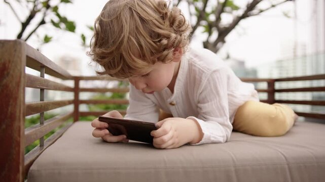 A 4-year-old curly-haired boy is completely absorbed playing games on his phone during a calm travel break. He comments on everything he sees, fully focused on the screen