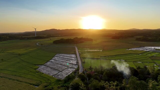 Aerial drone view of a wind turbine beside a rural road cutting through flat patchwork green and fallow rice fields at golden sunset, San Lorenzo Wind Farm, Guimaras, Philippines.