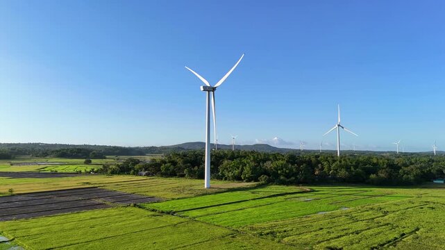 Aerial drone view of multiple white wind turbines rising above flat green and flooded rice paddies under a clear blue sky, San Lorenzo Wind Farm, Guimaras, Philippines.
