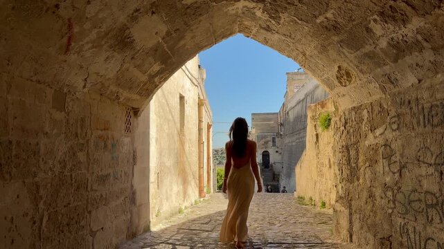 Rear View of Elegant Woman Walking Through Ancient Stone Archway in Sassi di Matera, UNESCO World Heritage Site, Basilicata, Italy, into Sunlit Historic Street in Cinematic Slow Motion