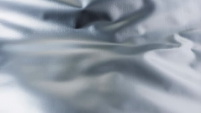 Slow motion closeup pan of silver satin fabric rippling and folding on bed linen in interior studio with subtle reflective texture and soft drape movement