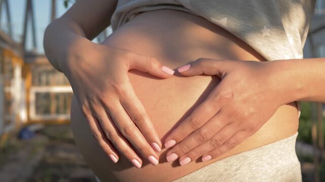 Future mother caressing pregnant belly and making heart shape with their hands. New life, motherhood and family concepts expressed in a loving and tender gesture outdoors