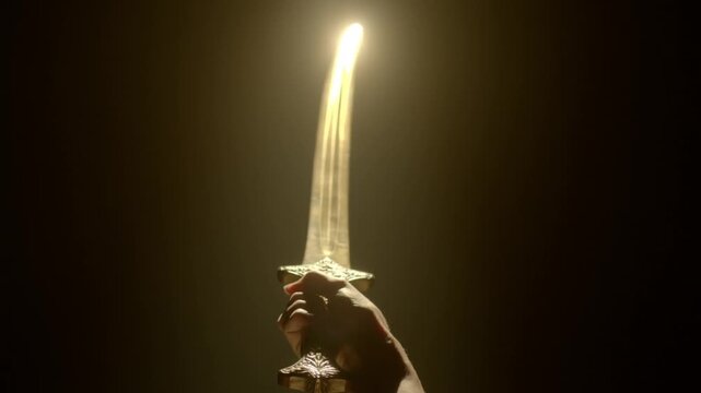 Slow motion tracking tilt of hand raising sword into spotlight on dark studio stage, close up ceremonial reveal of gleaming blade and hilt