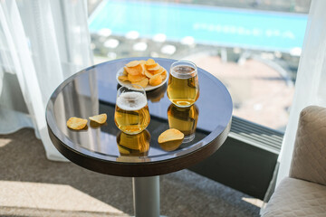 Obraz premium Two glasses of cold beer and potato chips on reflective table overlooking a resort pool. Bright sunny concept of travel, leisure and summer vacation relaxation in hotel room
