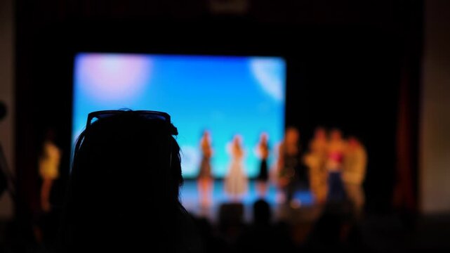 Spectators watch a fashion show on stage in a dark hall. Silhouette of a young woman in the foreground and blurred figures of models in the spotlights