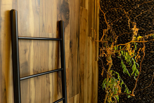 Close-up of a modern black metal ladder towel rail against a wooden wall. Stylish eco bathroom interior detail with decorative dry tree branches on a black pebble stone background.