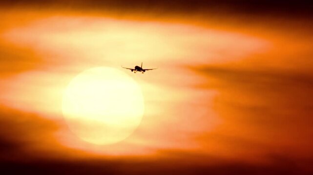 Passenger jet silhouette flying across a massive glowing sunset sun. Warm orange sky with hazy clouds during dusk, captured with extreme telephoto zoom lens.