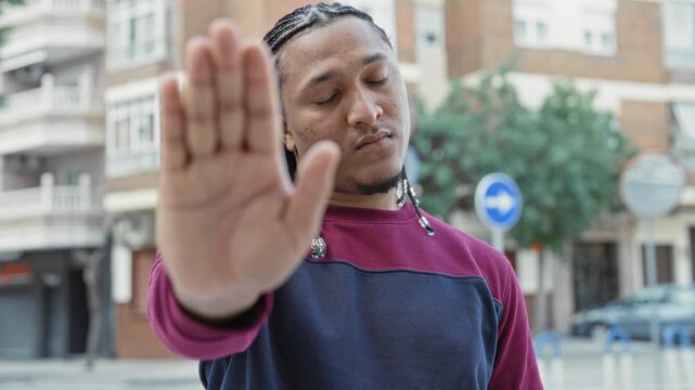 Man showing outstretched palm stop gesture on urban street with braided hair and sweatshirt; boundary defiance calm.