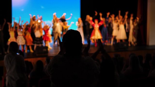 Spectators applaud the children's dance group during their final bow on stage. Silhouette of a person in the audience against the backdrop of a colorful performance