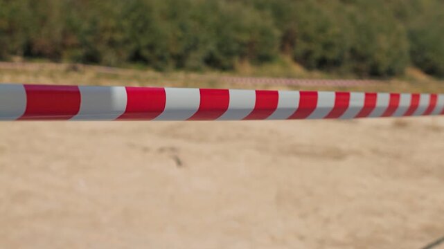 Red and white striped barrier tape stretched horizontally across sandy beach with blurred green vegetation background. Closeup view of safety warning tape marking restricted area
