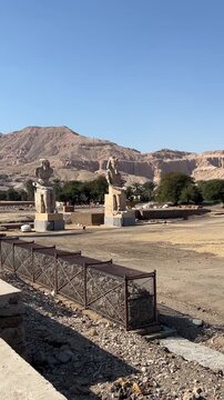 Ancient Colossi of Memnon, statues of Pharaoh Amenhotep III in Luxor, Egypt. Majestic historical landmarks set against a backdrop of desert mountains under clear blue skies. 
