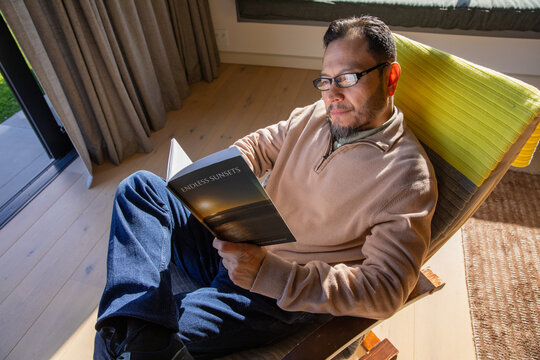 Asian man reclining in chair at home near glass door, wearing glasses and reading ENDLESS SUNSETS