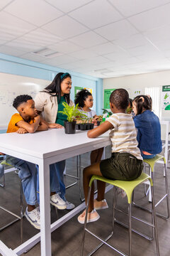 Teacher guiding four school-age children in classroom at white table, examining seedlings