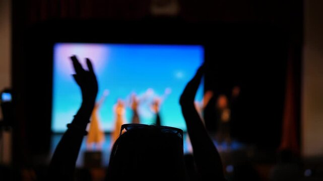 A spectator applauds the dancers on stage during the final performance in a concert hall. Silhouette of a person clapping their hands against the background of a bright screen
