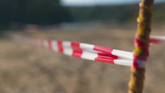 Red and white striped safety tape tied to yellow rebar flutters in the wind. Blurred background sandy ground and green hills. Safety warning concept. Shallow depth of field. Natural light.