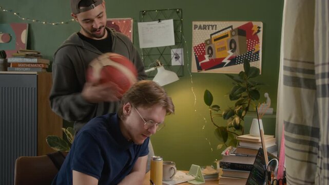 Caucasian young male studying at desk while roommate distracts him with basketball, asking to borrow money, handing over cash. Student lifestyle, roommate relationship, financial struggle ads