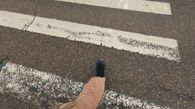 A first-person view of feet walking along markings of a worn-out pedestrian crossing. Cracks in asphalt indicate weathering. An urban street scene, focusing on road markings and pedestrian movement