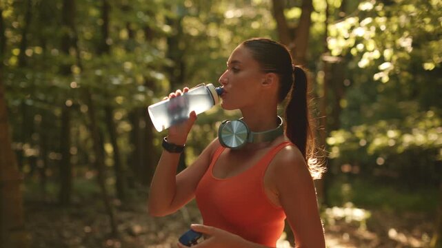 Woman standing in sunny forest and drinking water from bottle, taking break from jogging, staying hydrated throughout her activity, 4K, cinematic footage